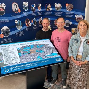 Three people smile beside a large interactive city map touchscreen in a blue timeline exhibit.