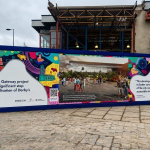 Colorful bannered construction fence with quotes about Derby's city centre and a photo of people in a plaza near a modern building under construction.