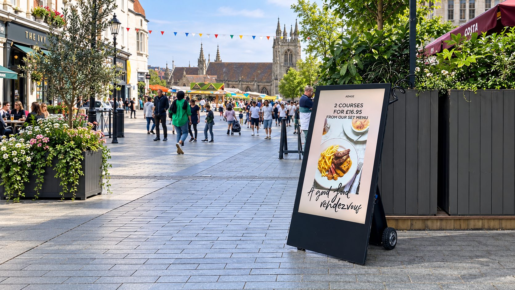 Busy pedestrian street with cafes and a flower-filled planter; a sandwich board advertises 2 courses for £16.95 from the set menu.