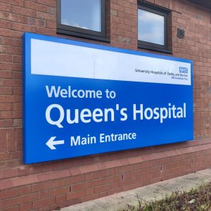 Blue sign on a brick wall reads "Welcome to Queen's Hospital, Main Entrance"