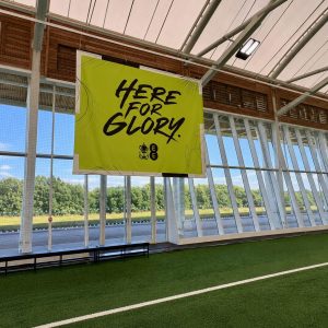 Bright lime banner reading 'Here for Glory' hanging inside a glass-walled sports hall with artificial turf and benches nearby.