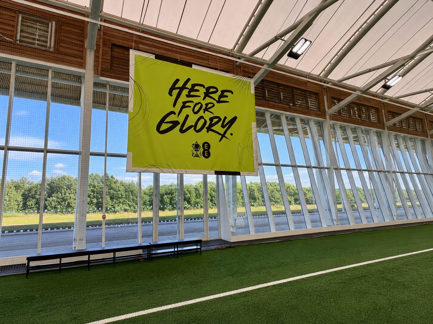 Bright lime banner reading 'Here for Glory' hanging inside a glass-walled sports hall with artificial turf and benches nearby.