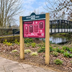Park information sign reading 'Burton Upon Trent' with a pink exhibit board near a river and iron bridge in a leaf-strewn garden path area.