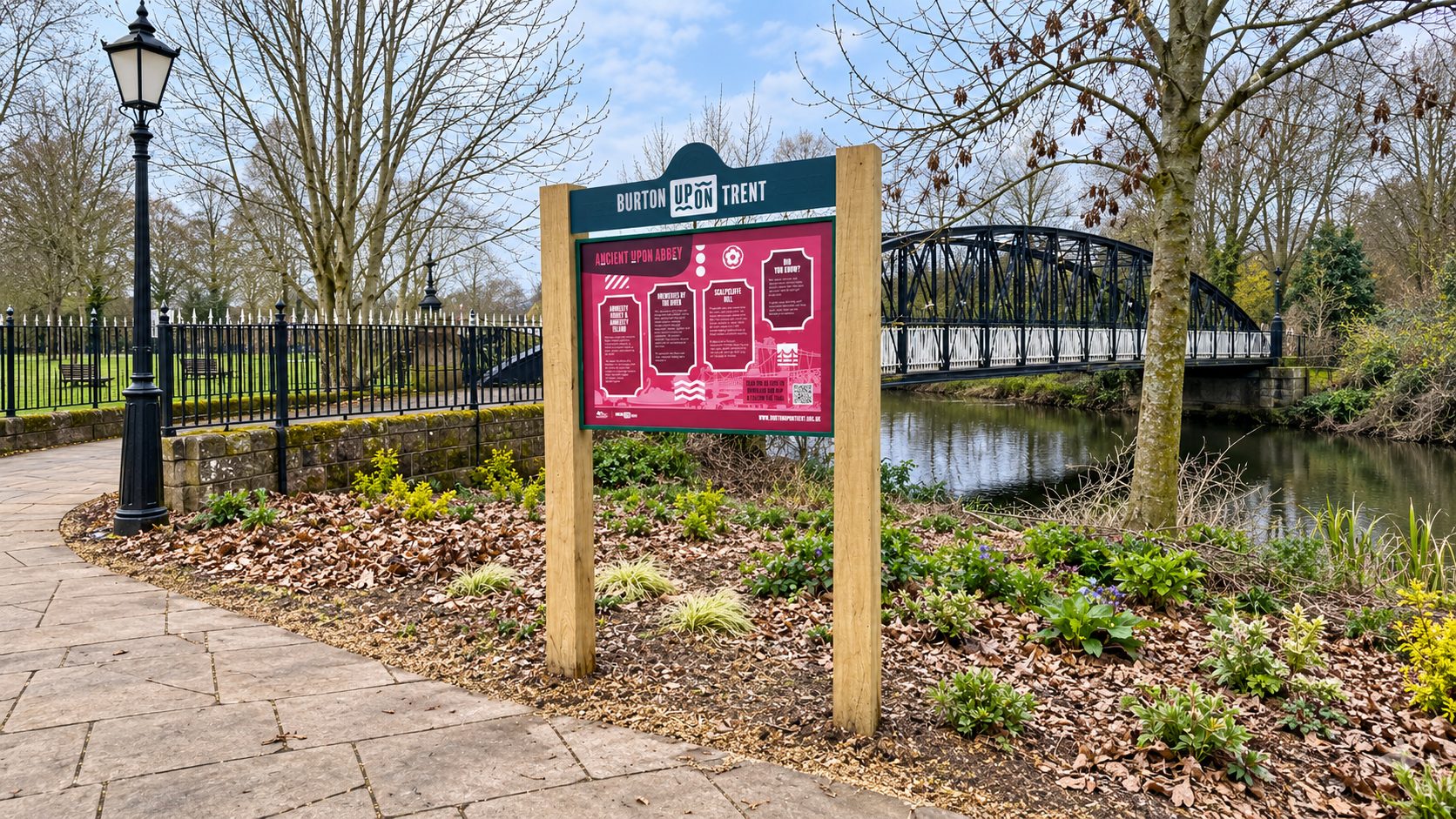 Park information sign reading 'Burton Upon Trent' with a pink exhibit board near a river and iron bridge in a leaf-strewn garden path area.