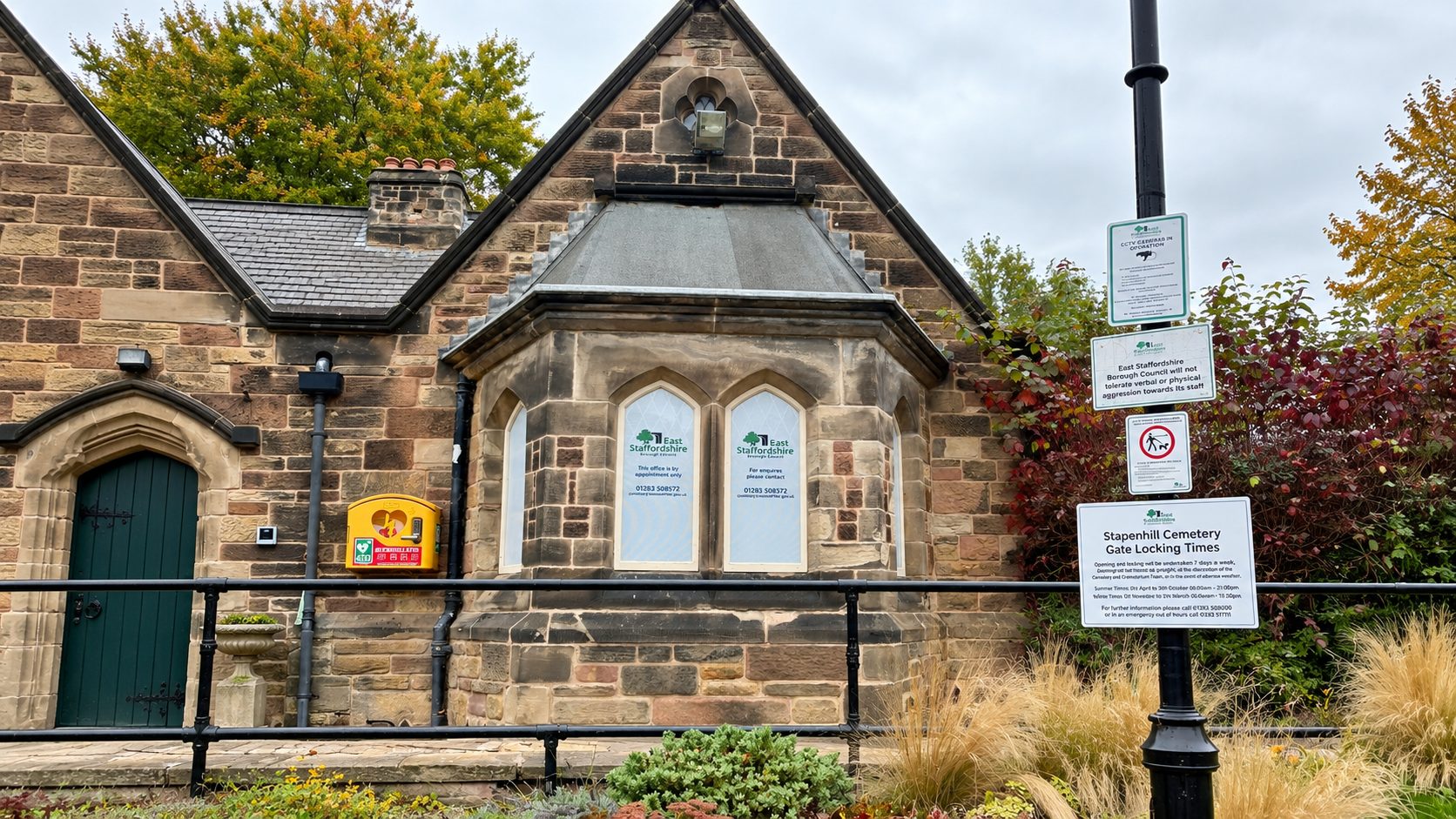 Stone chapel-like building with arched windows and a bright yellow mailbox on the wall, black railing in front, and a tall signpost with multiple notices nearby; autumn trees in the background.