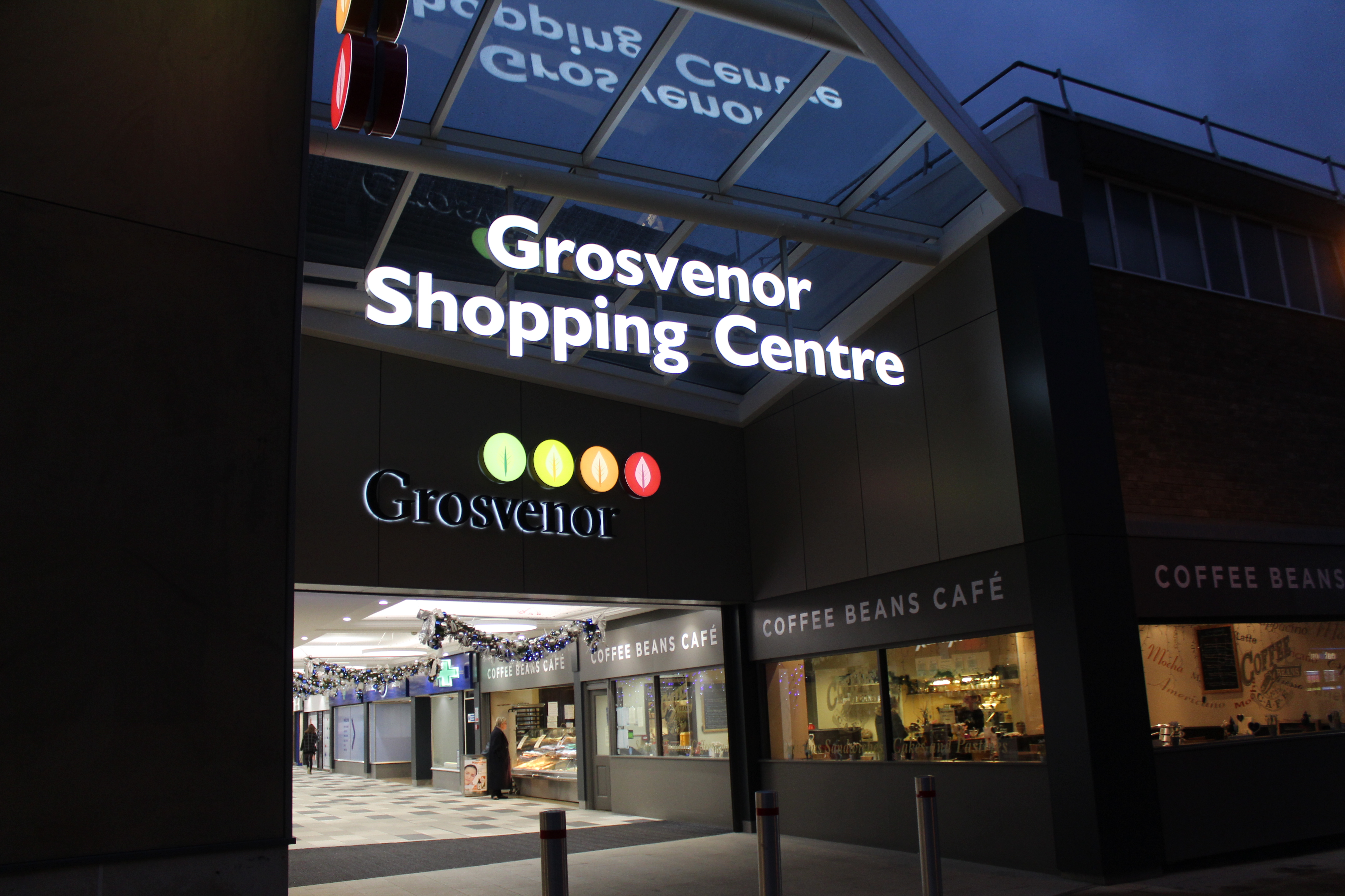 Night exterior of Grosvenor Shopping Centre with an illuminated sign and colorful circular logos under a glass canopy.