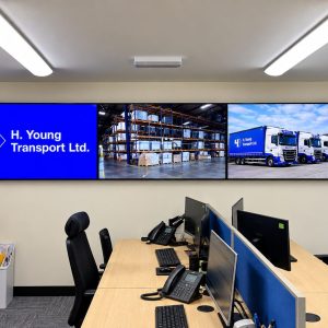 Office monitoring room with a three-panel wall display showing H. Young Transport branding, warehouse shelves, and blue-truck fleet set against beige walls.