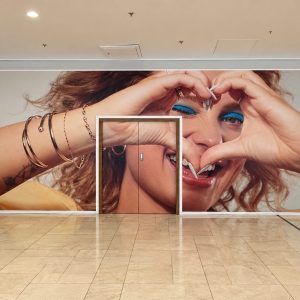 Large wall mural of a smiling woman making a heart shape with her hands around her eyes; double doors centered in the mural with red side panels.