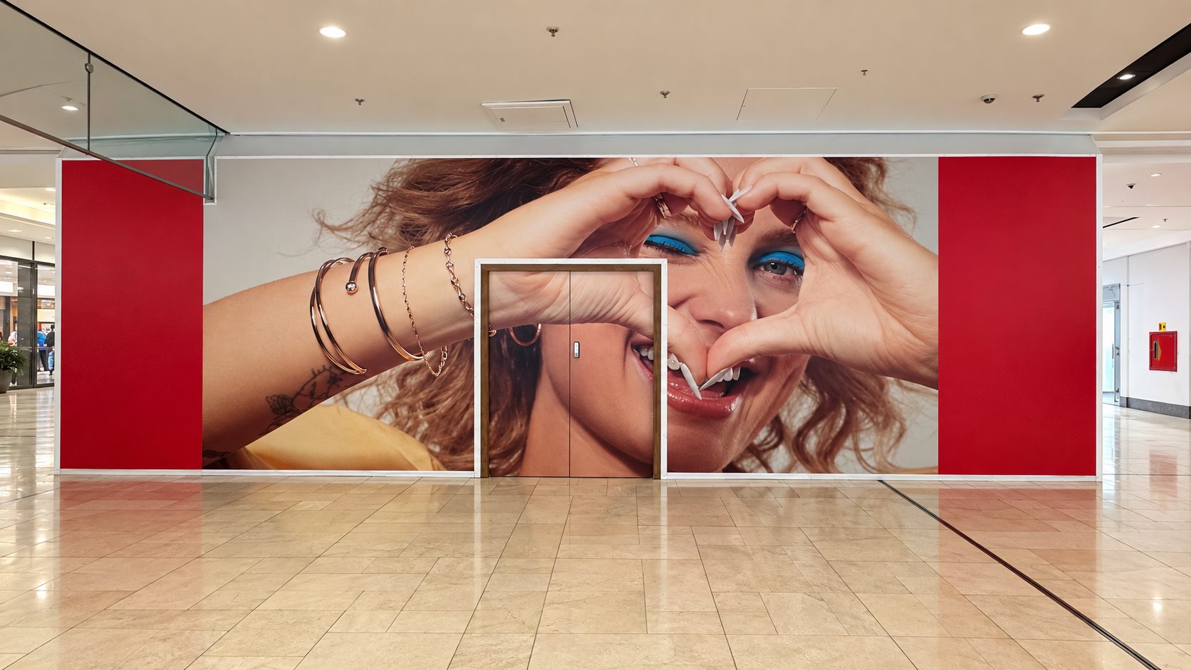 Large wall mural of a smiling woman making a heart shape with her hands around her eyes; double doors centered in the mural with red side panels.