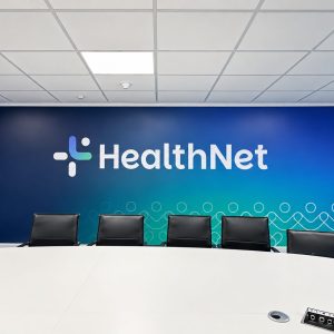 Conference room with a long white table and black chairs facing a blue gradient wall featuring the HealthNet logo, plant in corner.