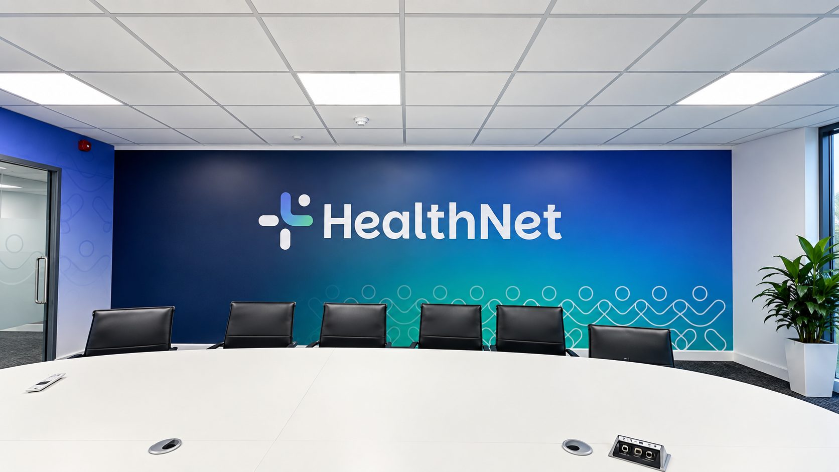 Conference room with a long white table and black chairs facing a blue gradient wall featuring the HealthNet logo, plant in corner.