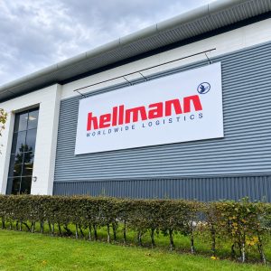 Exterior view of a gray industrial warehouse with a large white sign reading 'Hellmann Worldwide Logistics' in red letters.