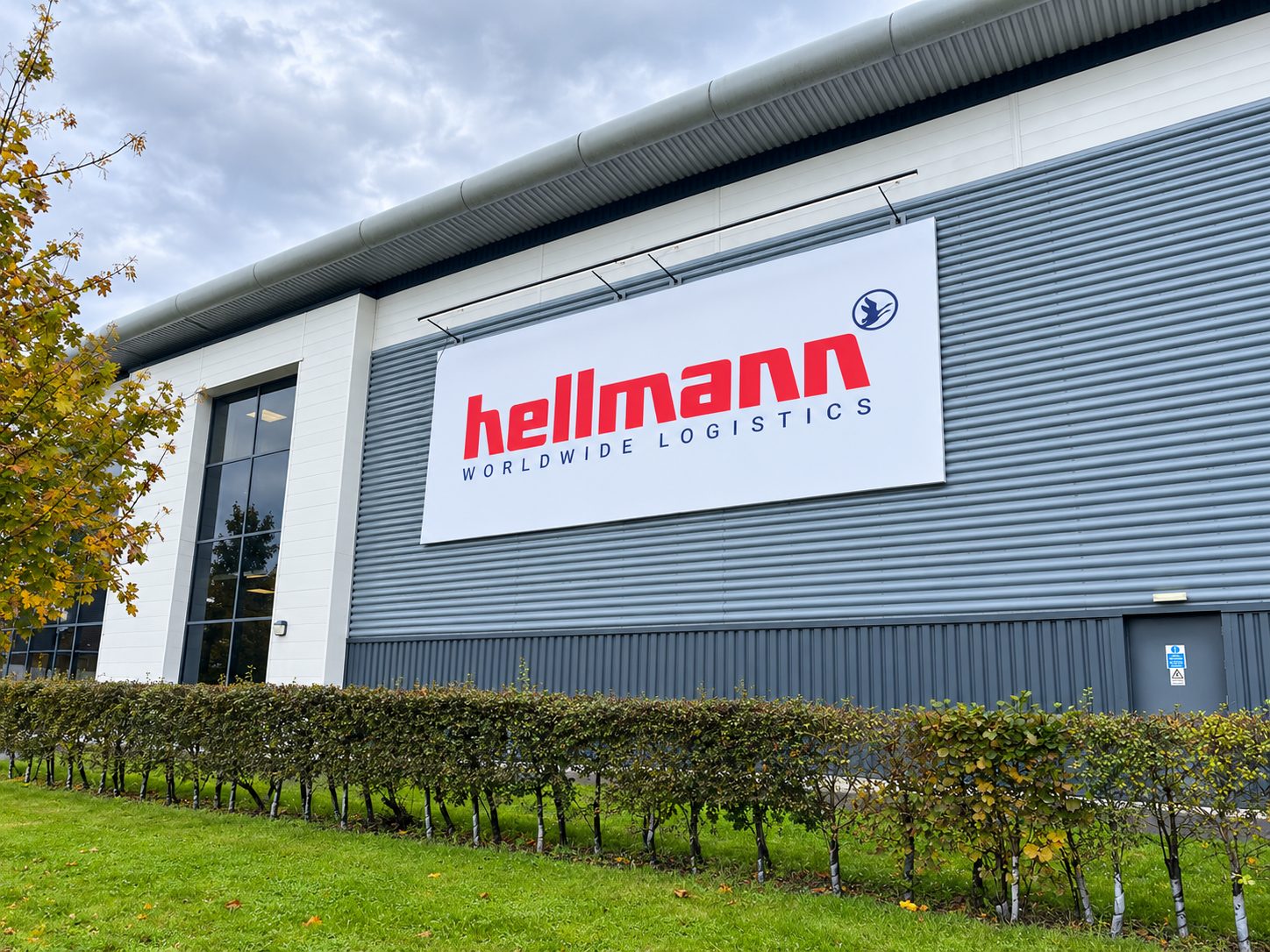 Exterior view of a gray industrial warehouse with a large white sign reading 'Hellmann Worldwide Logistics' in red letters.