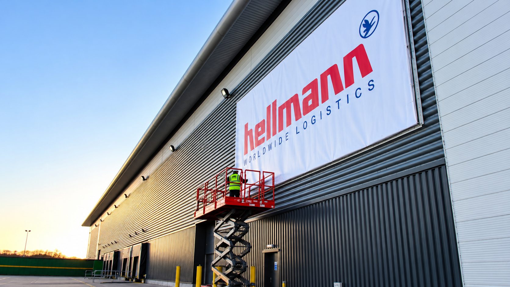 Industrial building exterior with a large 'hellmann Worldwide Logistics' banner being installed; a worker on a red scissor lift is affixed under the banner at dusk/dawn light.
