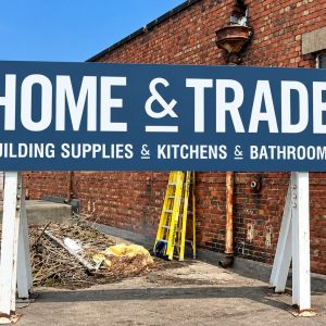 Blue sign says 'HOME & TRADE' with subtitle 'BUILDING SUPPLIES & KITCHENS & BATHROOMS' at a brick building site; a worker in a high-vis vest stands nearby.