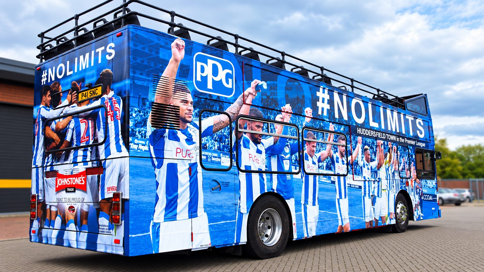 Team bus wrapped in blue with Huddersfield Town players celebrating, featuring the hashtag #NOLIMITS and sponsor logos.