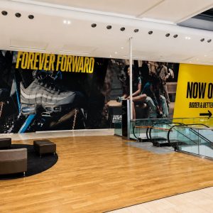 Indoor shopping mall atrium with a large sneaker ad reading 'FOREVER FORWARD' and a bright yellow 'NOW OPEN' JD banner beside an escalator.