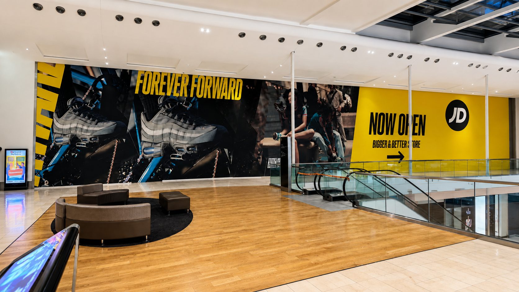Indoor shopping mall atrium with a large sneaker ad reading 'FOREVER FORWARD' and a bright yellow 'NOW OPEN' JD banner beside an escalator.