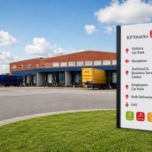 KP Snacks distribution center with a tall white sign listing directions to car parks, reception, and deliveries; loading bays and blue warehouse in the background.