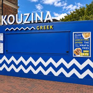 Blue Kouzina Greek food stand with large white sign, blue facade, and white zigzag pattern along the bottom; menu board on the left and a promotional poster featuring a lunch deal on the right.