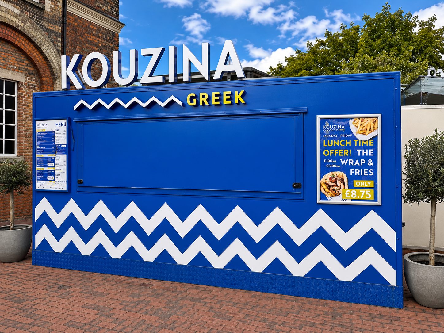 Blue Kouzina Greek food stand with large white sign, blue facade, and white zigzag pattern along the bottom; menu board on the left and a promotional poster featuring a lunch deal on the right.