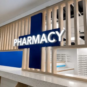Modern pharmacy counter with a bright white 'PHARMACY' sign mounted on a blue cross, behind a light wood slat divider.