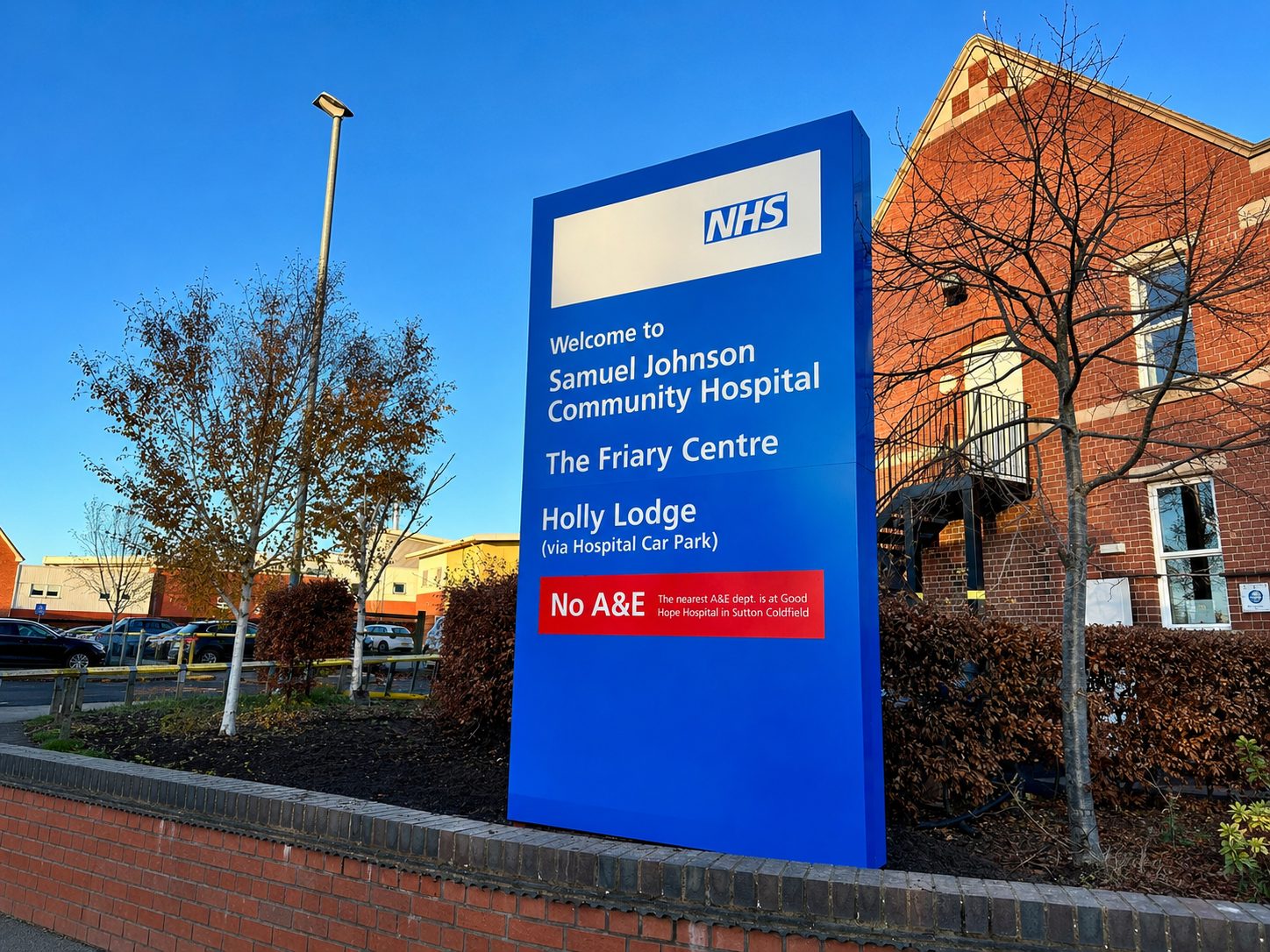 Blue NHS sign outside Samuel Johnson Community Hospital listing The Friary Centre and Holly Lodge with 'No A&E' notice (informational)