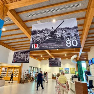 Indoor museum lobby with large black-and-white banners reading VJ Day 80 and featuring soldiers on an anti-aircraft gun, Royal British Legion logo visible. Visitors move about near a merchandise shop and reception desks under wooden beams.