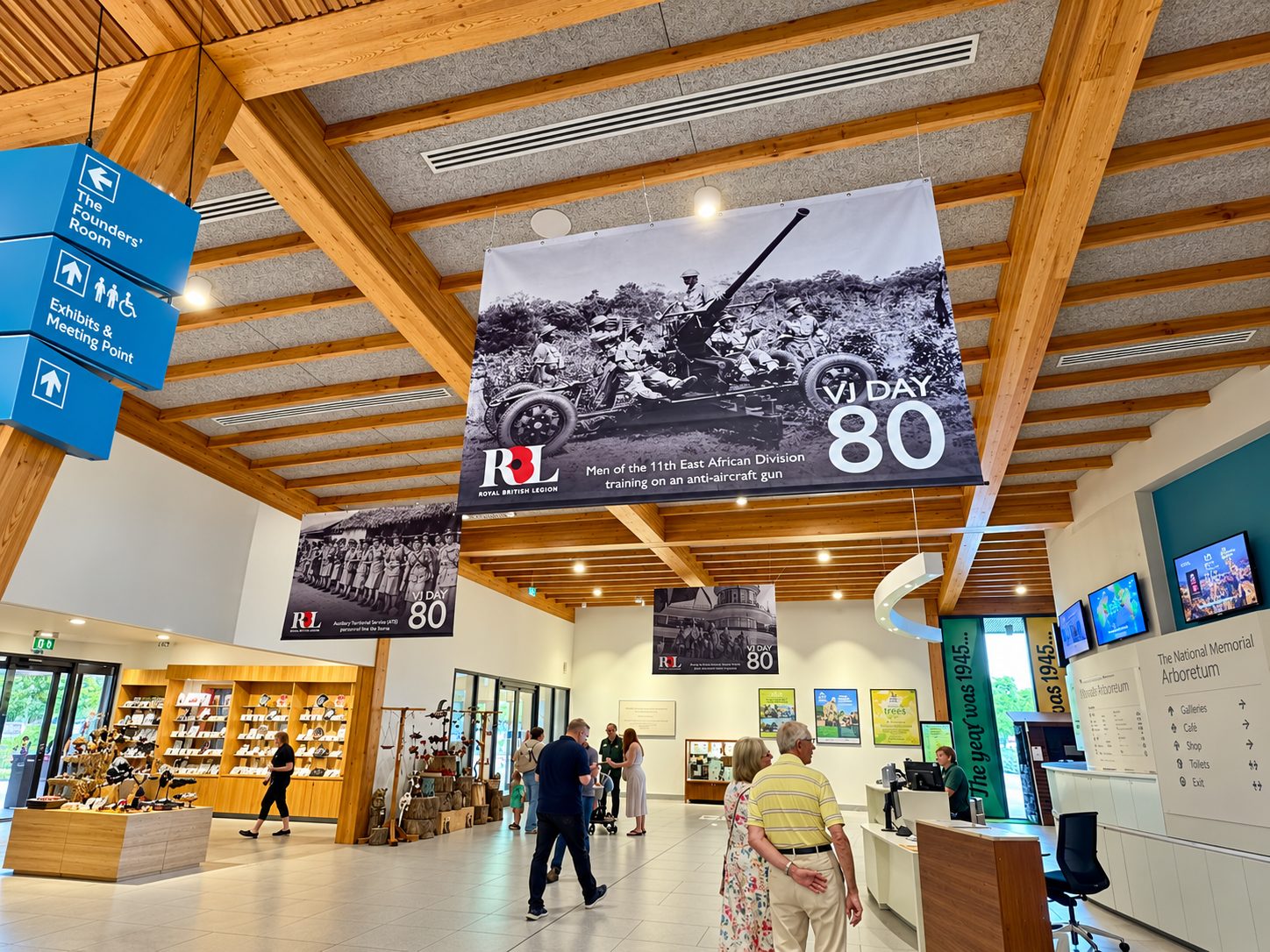Indoor museum lobby with large black-and-white banners reading VJ Day 80 and featuring soldiers on an anti-aircraft gun, Royal British Legion logo visible. Visitors move about near a merchandise shop and reception desks under wooden beams.