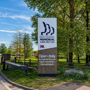 Sign for National Memorial Arboretum with slogan 'Open daily throughout the year' and 'Open from 10am' on a wood-textured base, trees and parking area in background