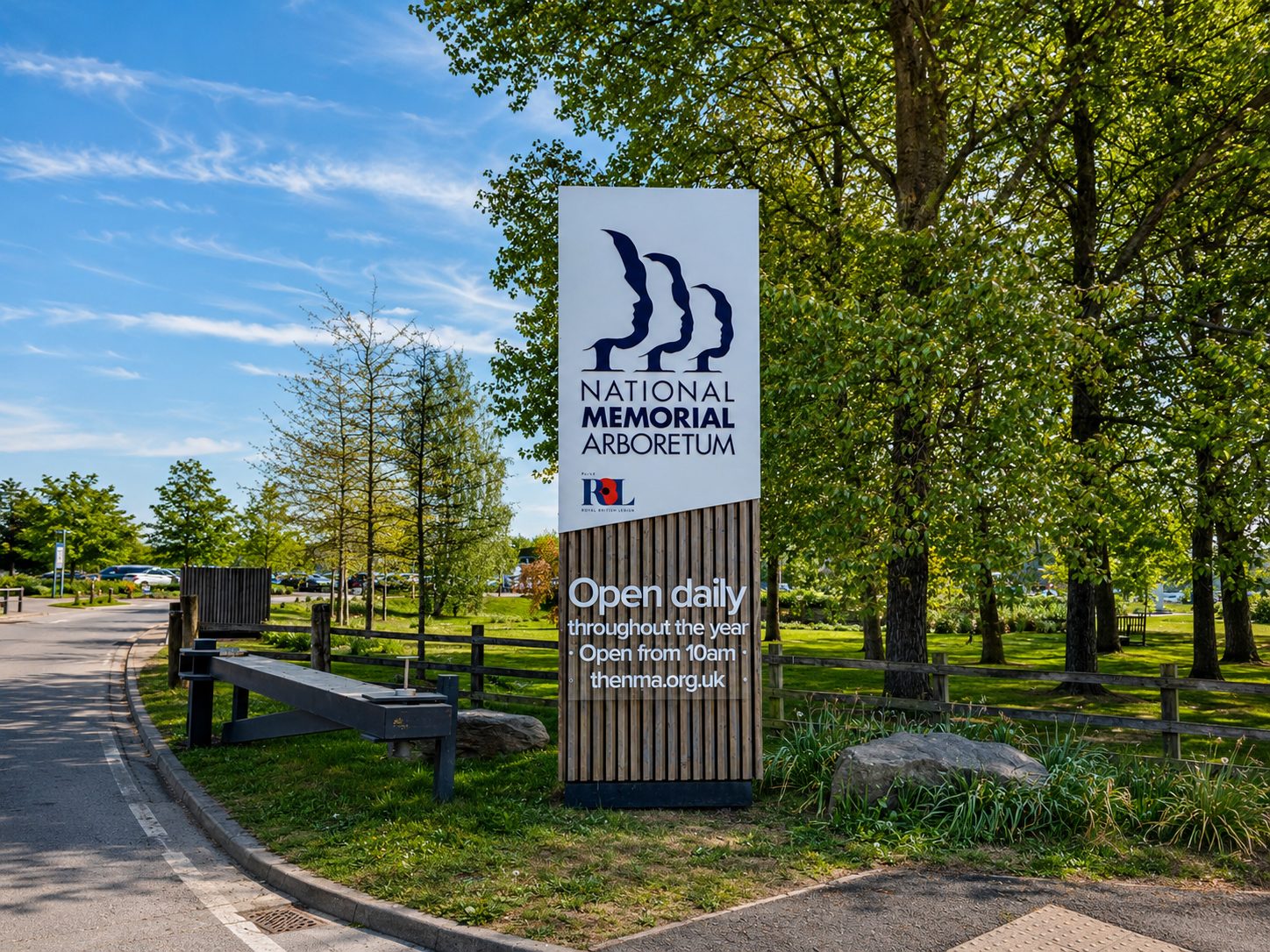 Sign for National Memorial Arboretum with slogan 'Open daily throughout the year' and 'Open from 10am' on a wood-textured base, trees and parking area in background