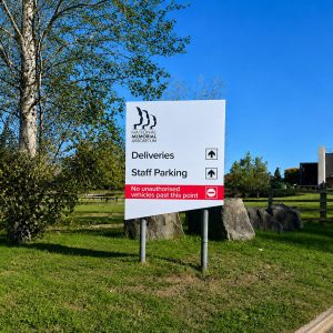 White sign for National Memorial Arboretum directs deliveries and staff parking; red strip warns no unauthorized vehicles past this point, with trees and a fence in the background.