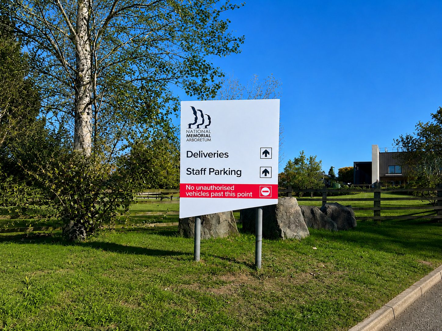 White sign for National Memorial Arboretum directs deliveries and staff parking; red strip warns no unauthorized vehicles past this point, with trees and a fence in the background.