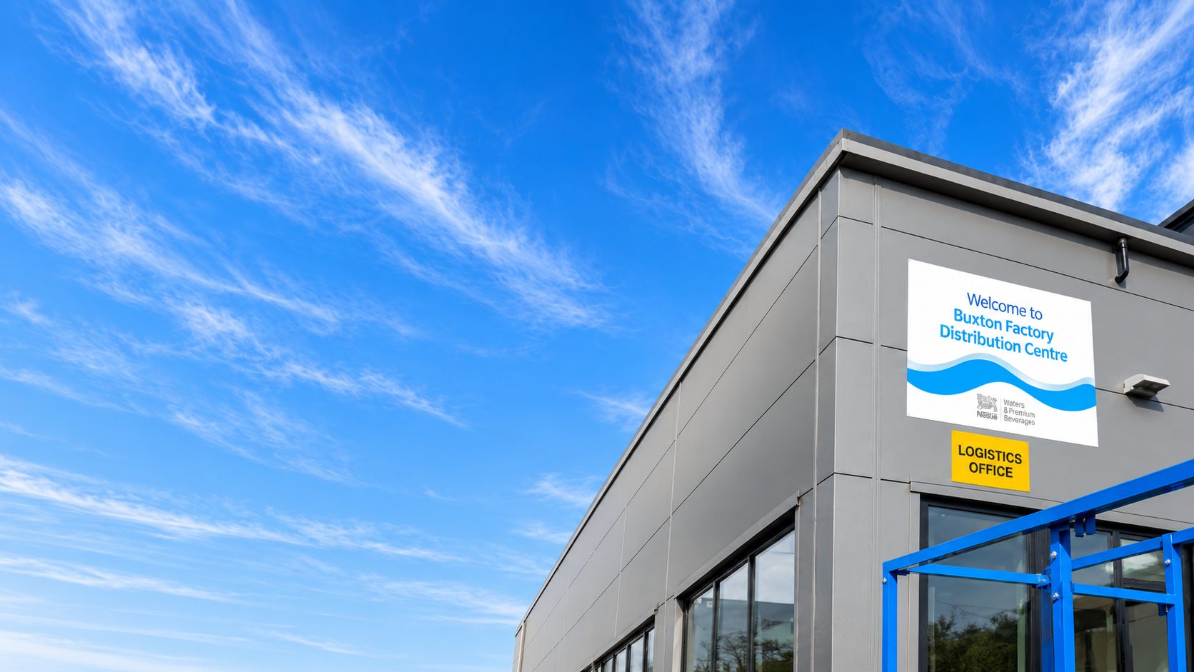 Factory corner with a large white welcome sign and a yellow logistics office sign on a gray building, blue sky above and blue framework in foreground.