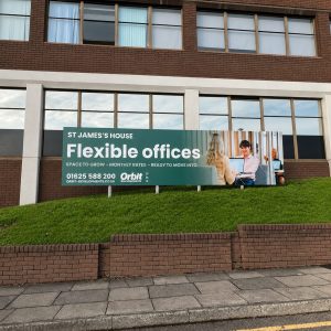 Green billboard on a grassy slope promoting 'Flexible offices' at St James's House with contact info, in front of a brick building