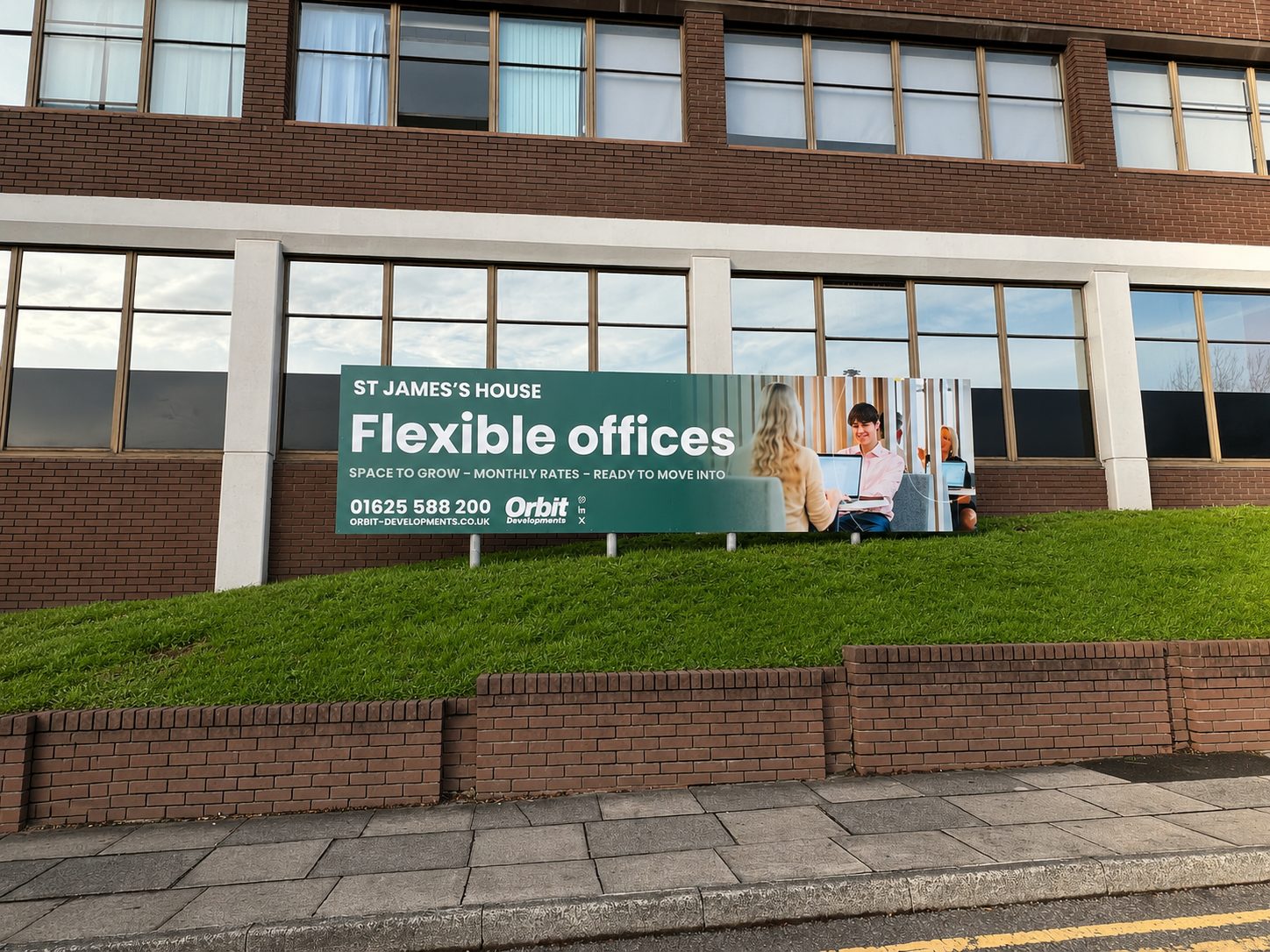 Green billboard on a grassy slope promoting 'Flexible offices' at St James's House with contact info, in front of a brick building