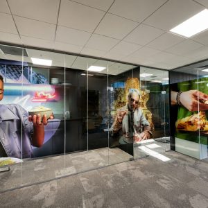 Colorful hallway mural featuring people enjoying food, visible behind glass walls in an office space.