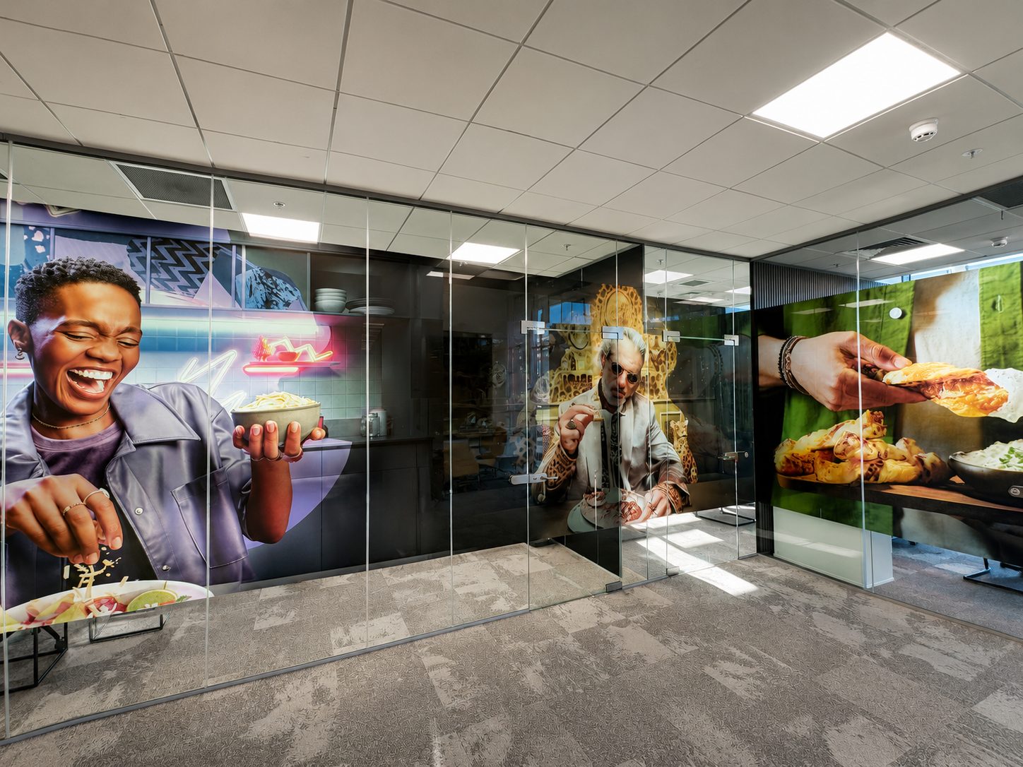 Colorful hallway mural featuring people enjoying food, visible behind glass walls in an office space.