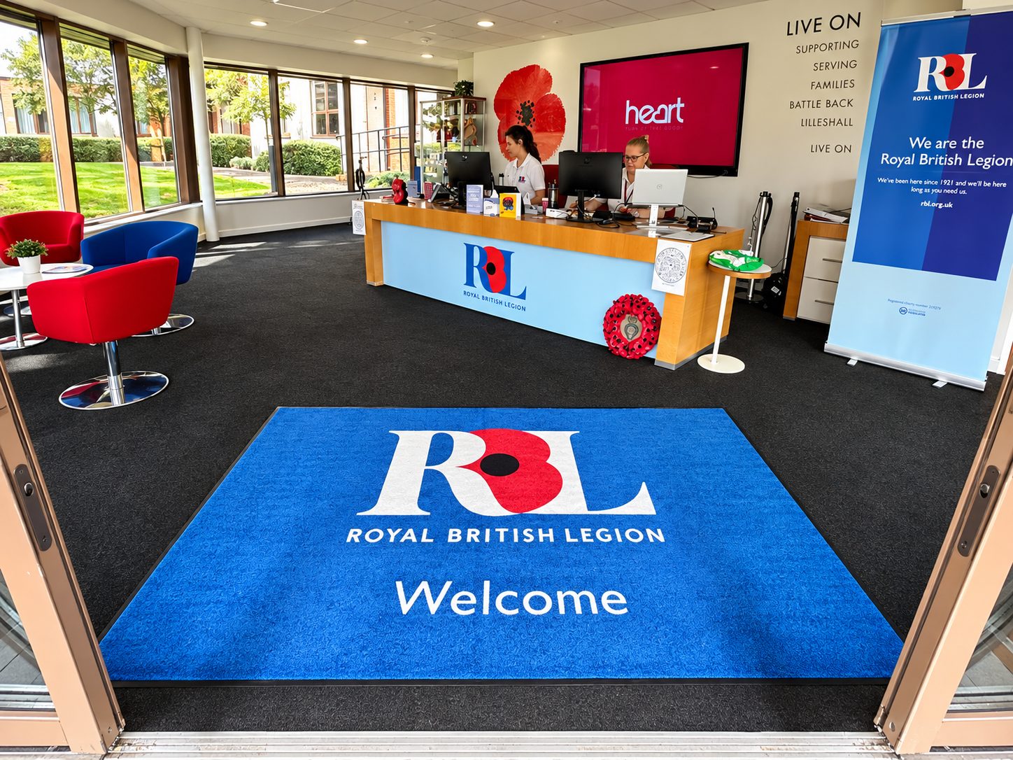 Blue welcome mat with the Royal British Legion logo and 'Welcome' at the doorway, leading into a reception area with staff at a desk.