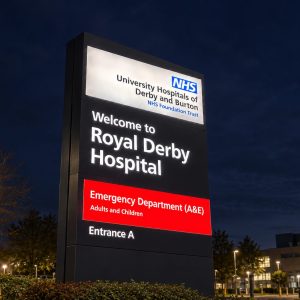 Illuminated hospital sign at night reading 'Welcome to Royal Derby Hospital' with red A&E panel, NHS logo visible.