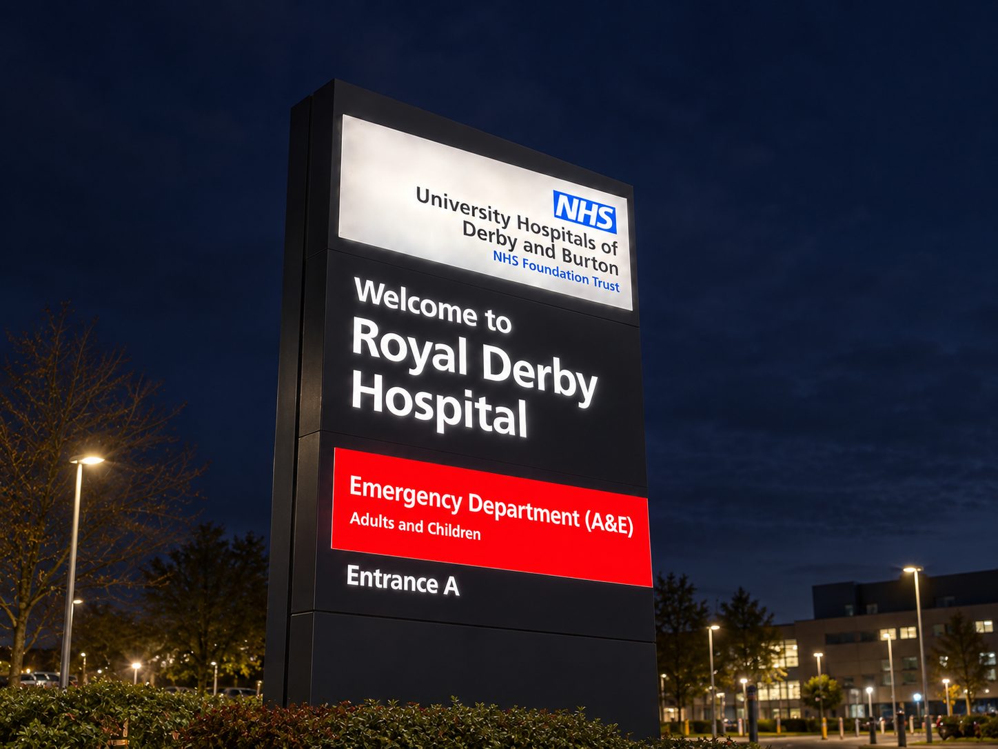 Illuminated hospital sign at night reading 'Welcome to Royal Derby Hospital' with red A&E panel, NHS logo visible.