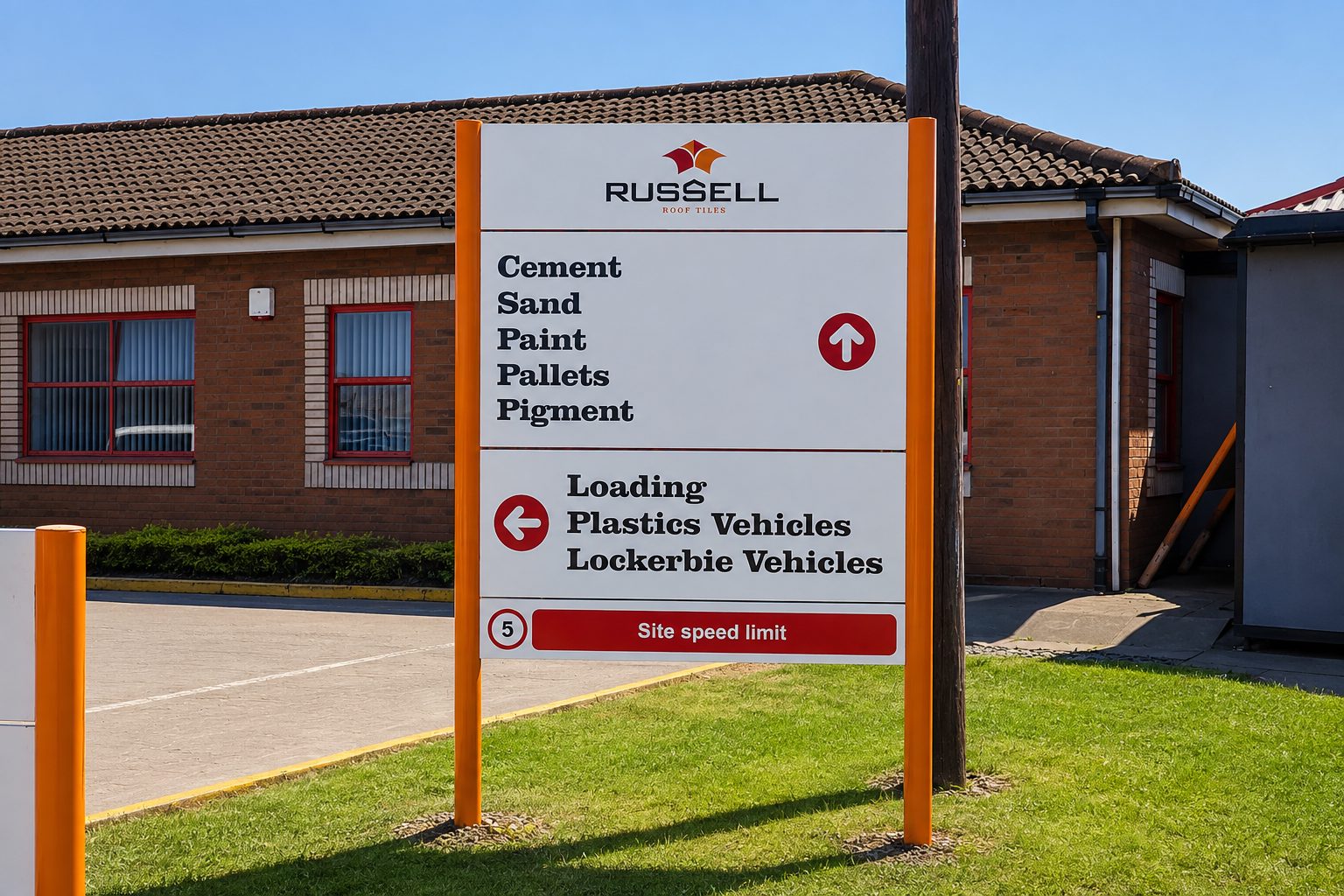 Large outdoor sign at a building materials site listing cement, sand, paint, pallets, pigment with directional arrows and a red 'site speed limit' bar; Russell Roof Tiles logo visible; brick building in background.