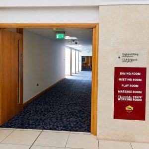 A doorway into a hallway with a blue-patterned carpet; wall sign listing dining, meeting, play, massage, and technical staff rooms on a red panel to the right.