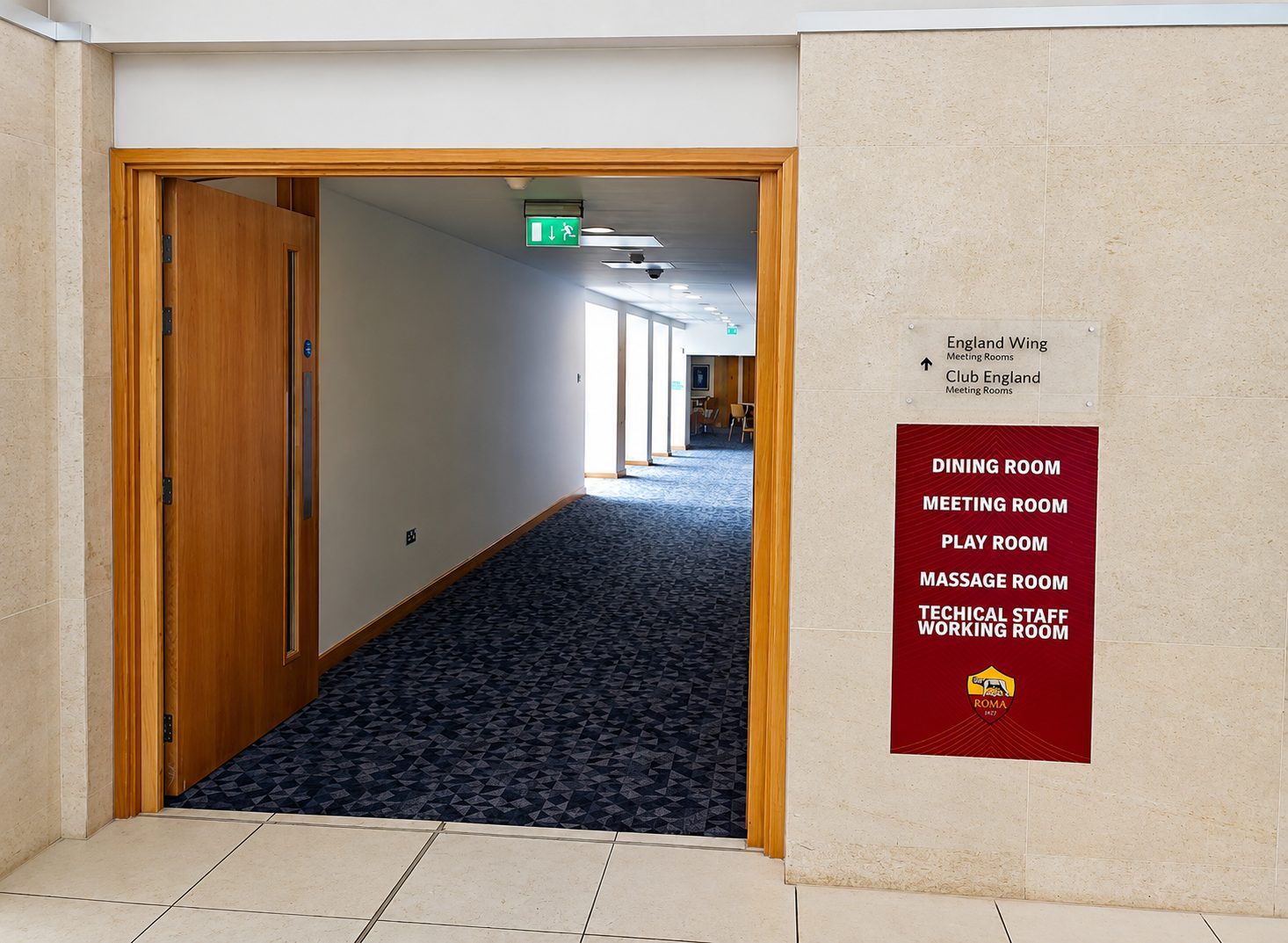 A doorway into a hallway with a blue-patterned carpet; wall sign listing dining, meeting, play, massage, and technical staff rooms on a red panel to the right.