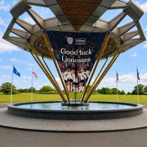 Large circular fountain under a modern geometric pavilion with a banner reading 'Good luck Lionesses' and flagpoles around the lawn.
