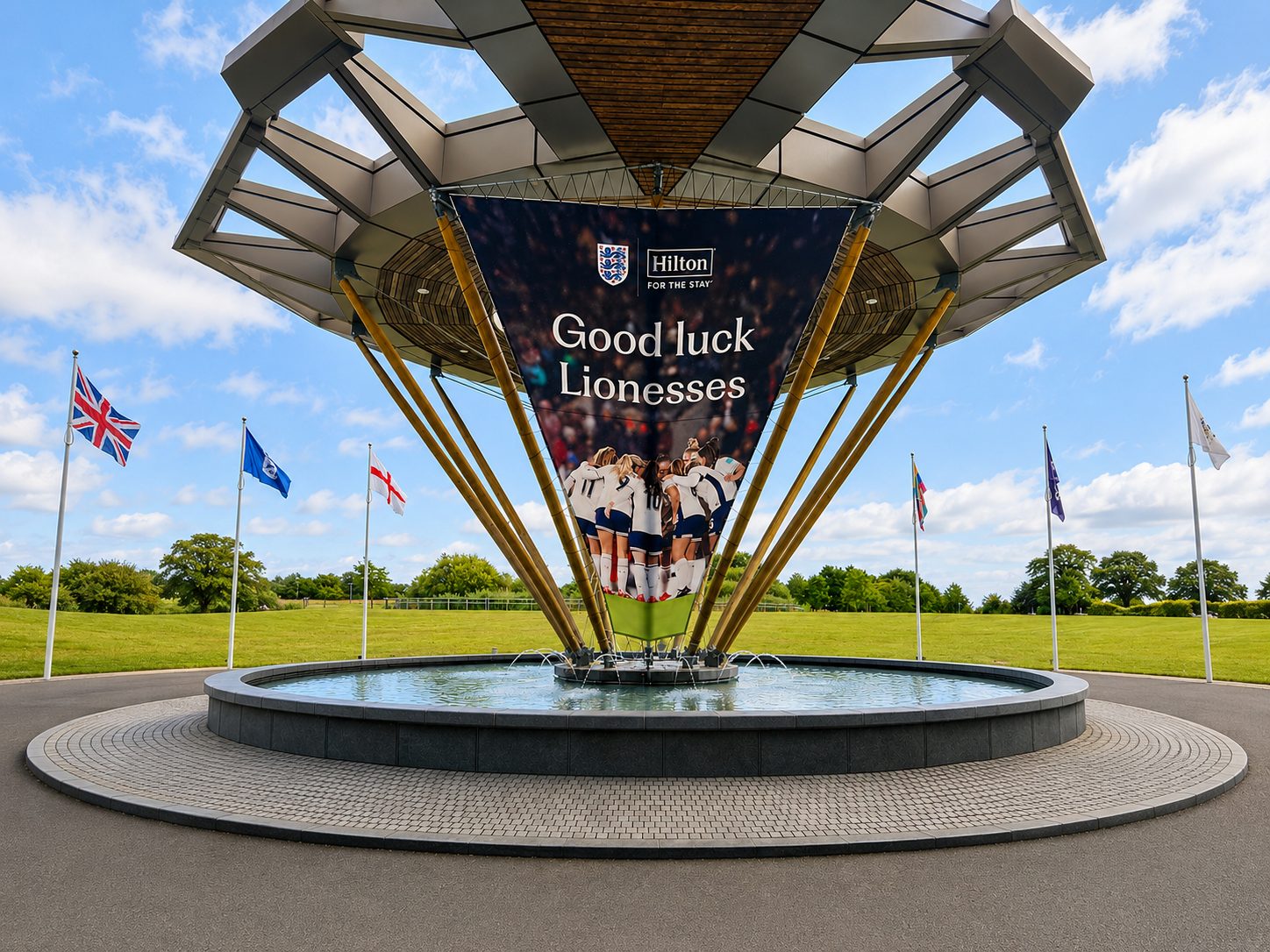 Large circular fountain under a modern geometric pavilion with a banner reading 'Good luck Lionesses' and flagpoles around the lawn.