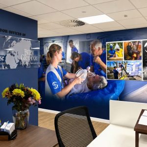 Reception area of a humanitarian charity office with blue wall signage: Samaritan's Purse and Billy Graham logos, plus a collage mural of relief images on the right panel.