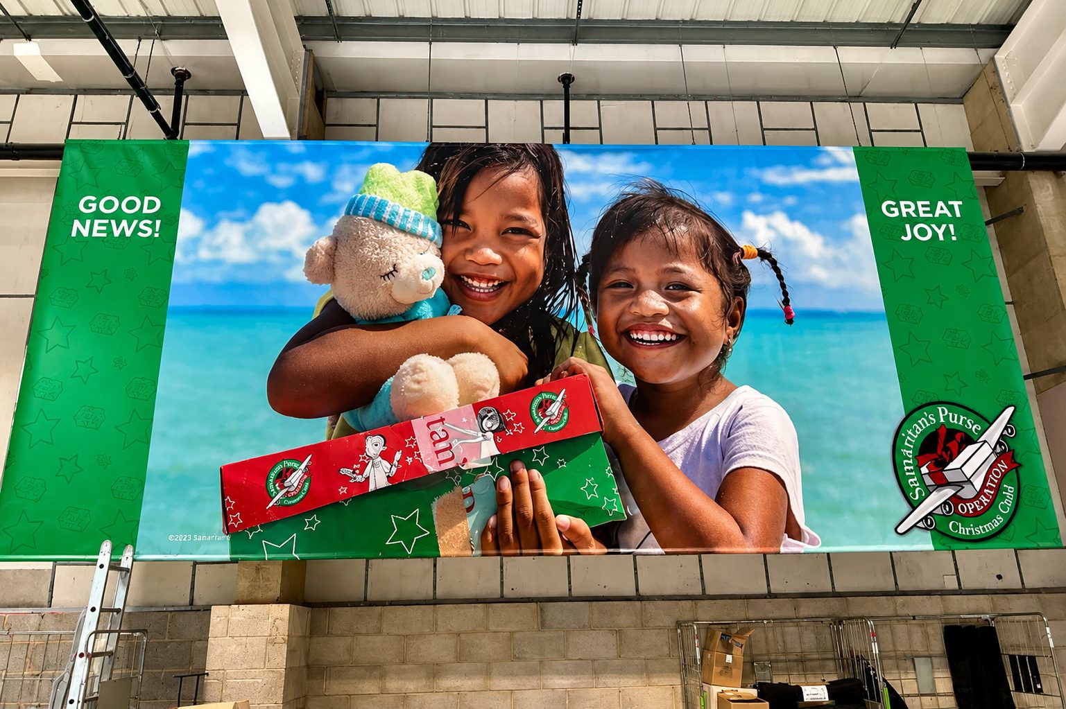 Two smiling children hug a teddy bear and a wrapped gift on a bright banner showing 'Good News!' and 'Great Joy!' by a beach scene within an indoor display