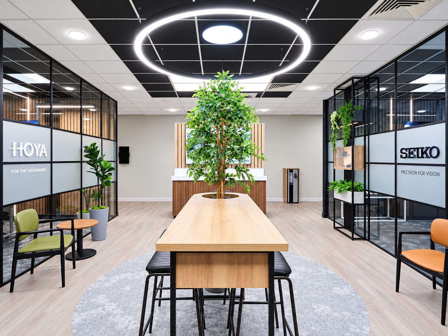 Modern office showroom with a central wooden table, a tall potted tree, and a circular ceiling light ring surrounded by glass partitions labeled Hoya and Seiko.