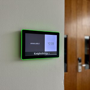 Wall-mounted tablet with a bright green bezel showing 'AVAILABLE' and 'Knightsbridge 1' beside a wooden door panelled wall.